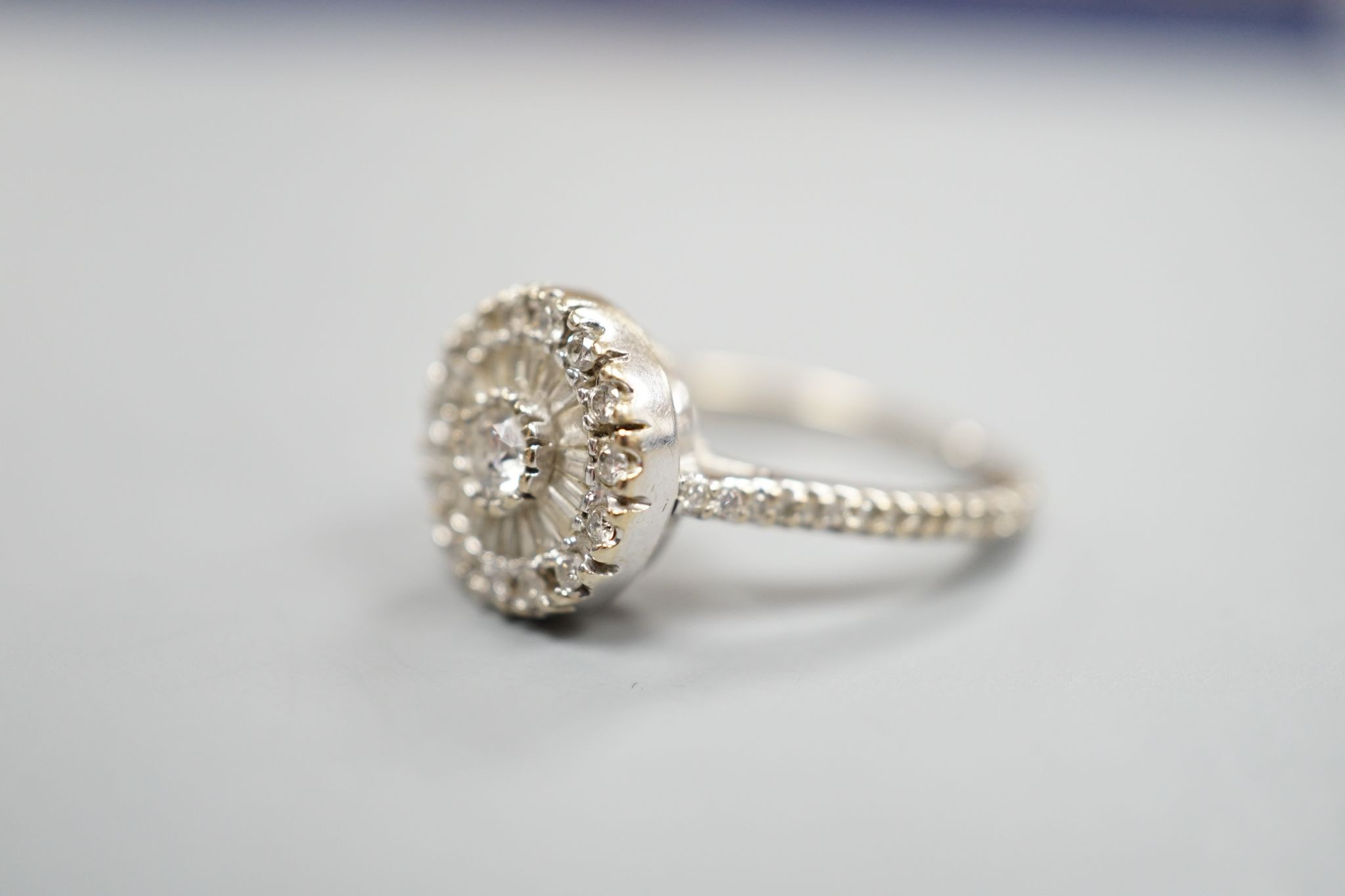 A modern white metal, round and trapeze cut diamond set target cluster ring, with diamond set shoulders, size N, gross weight 4.2 grams.